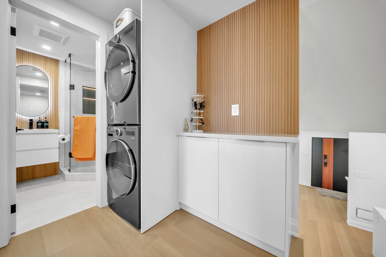 Bathroom with wood cabinetry, a glass shower, a round mirror and stacked laundry appliances.